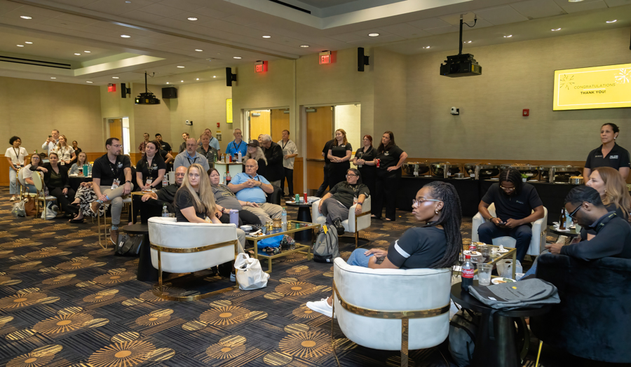 Group of people seated in a hotel conference room, some standing at the back, watching a presentation with a snack area along the wall.