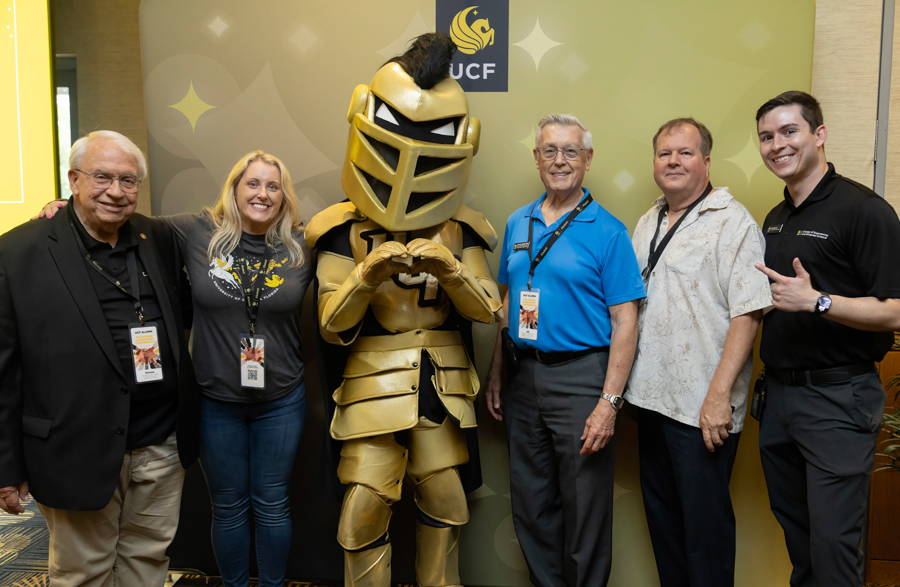 Six adults posing with a gold armored knight mascot in front of a UCF-branded green backdrop at an event.
