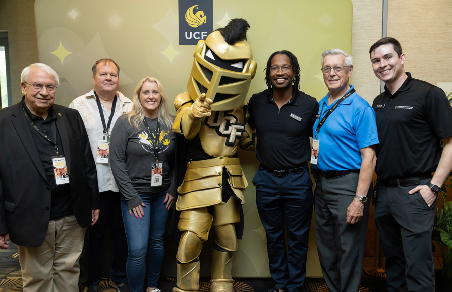 Seven people pose with a gold knight mascot in front of a UCF banner at an event, all smiling and wearing name badges.