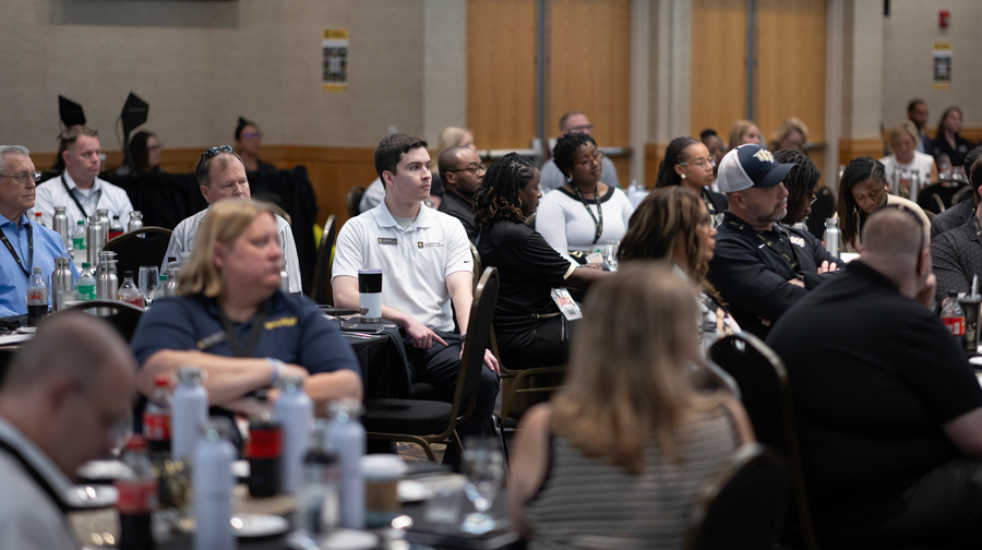 Group of adults seated at round tables in a conference room, listening to a presentation.