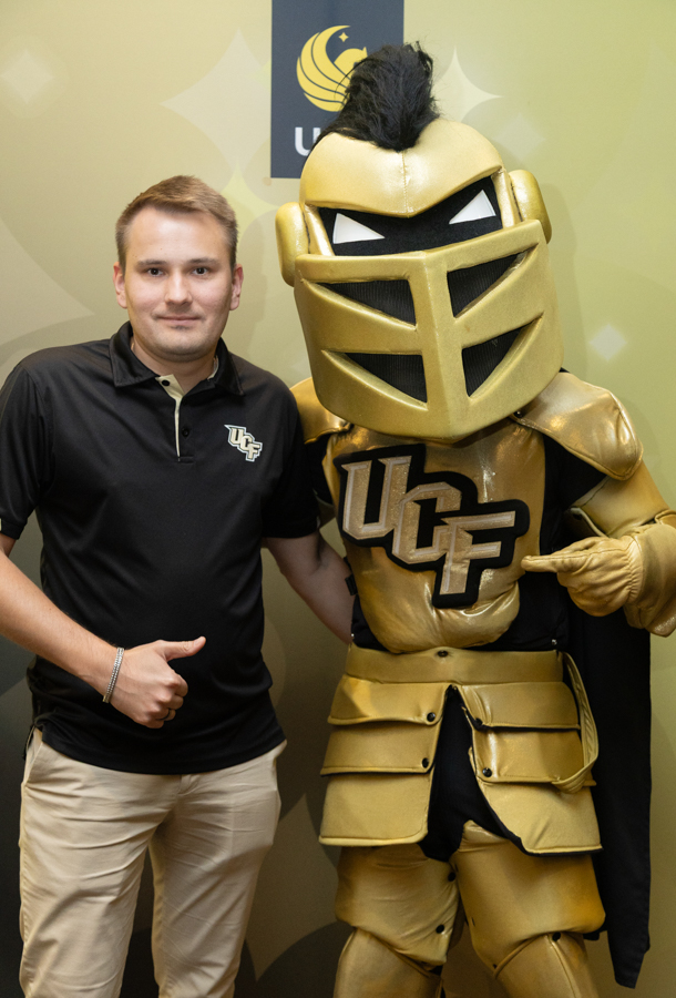 Man in a black polo with a UCF logo posing next to a gold knight mascot with UCF lettering on its chest.