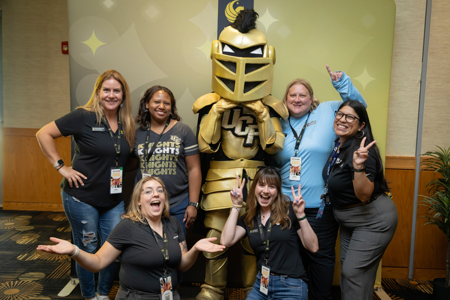 Six people—five women and a person in a gold knight mascot costume—pose and smile for a group photo indoors.