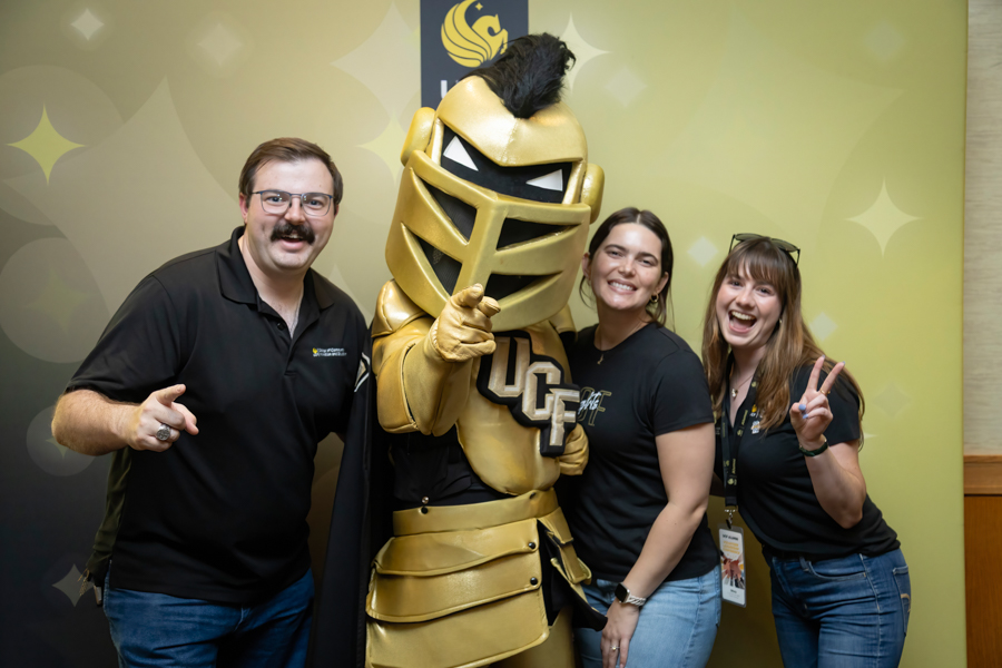Group of three people posing with a gold knight mascot in front of a lime-green backdrop, all smiling and making peace signs or thumbs-up.