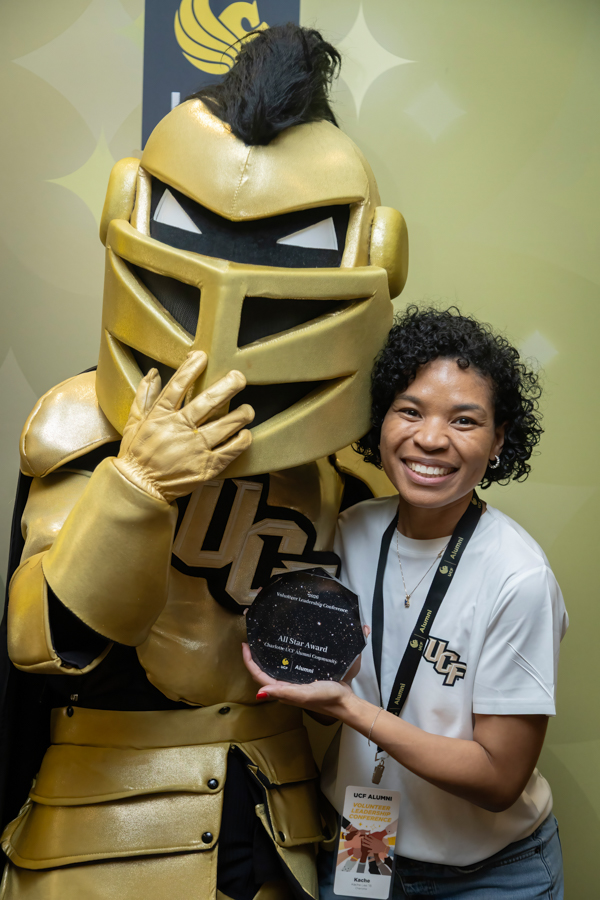 Smiling woman with a lanyard stands beside a performer in a large gold knight costume, holding a round award plaque.