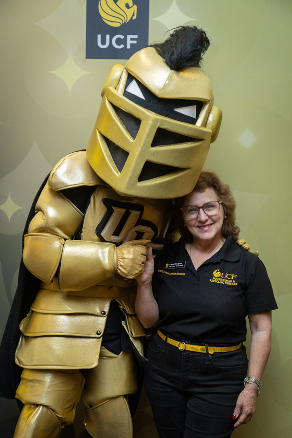 Woman smiling in a black UCF polo standing next to a person in shiny gold knight armor, posing for a photo with the UCF logo in the background.