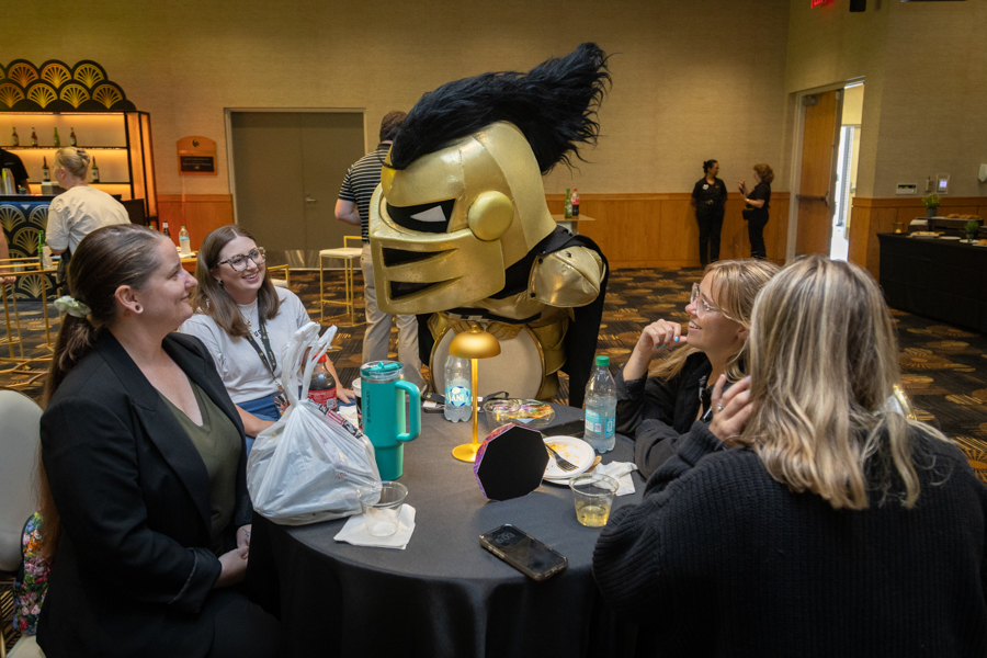 Four women sit around a round banquet table, talking with a large gold knight mascot with a black mohawk in a event space.