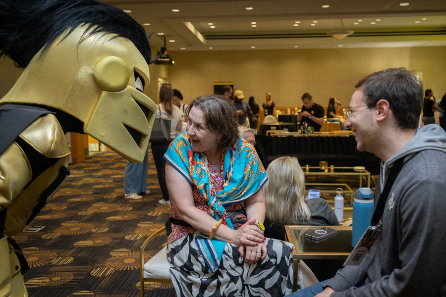 A person in a large gold mascot costume leans toward a seated woman in a bright blue scarf, in a conference hall with people in the background, smiling man nearby.