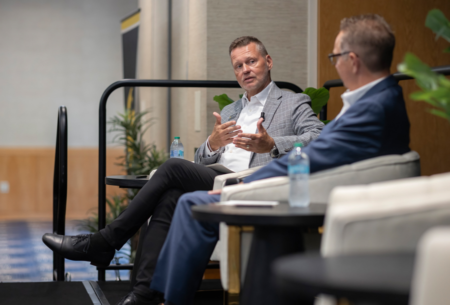 Two men in a business panel: one speaking with hand gestures while seated on a stage with water bottles nearby