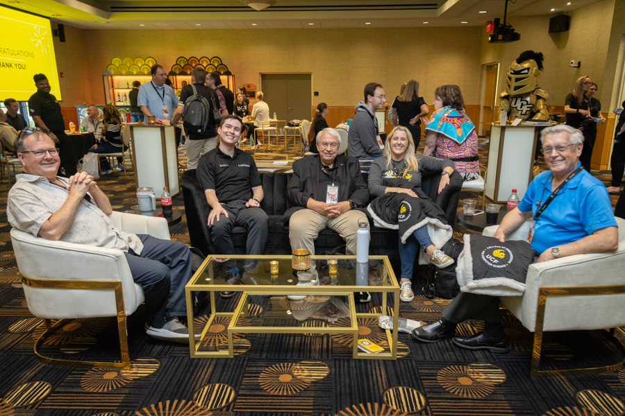 Six adults cheerfully pose for a casual group photo, seated on couches around a glass coffee table in a hotel lounge at a conference reception.