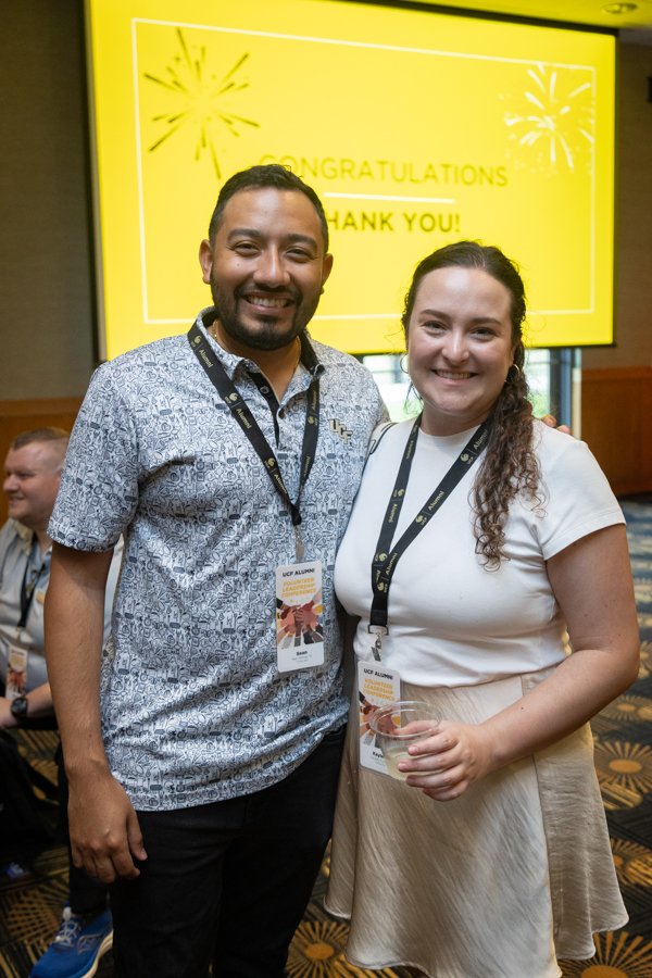 Two event attendees posing together and smiling, with a bright yellow screen reading 'CONGRATULATIONS' behind them.