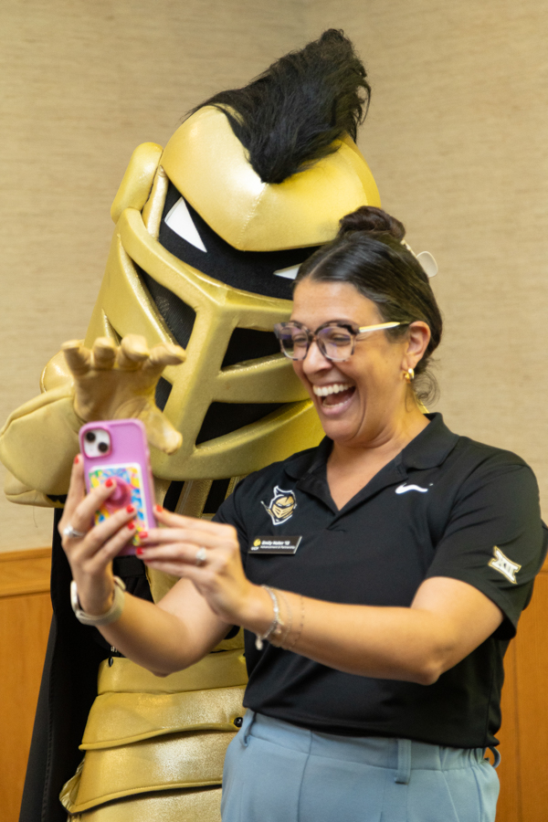 Woman in a black polo taking a selfie with a smiling gold knight mascot beside her.”,