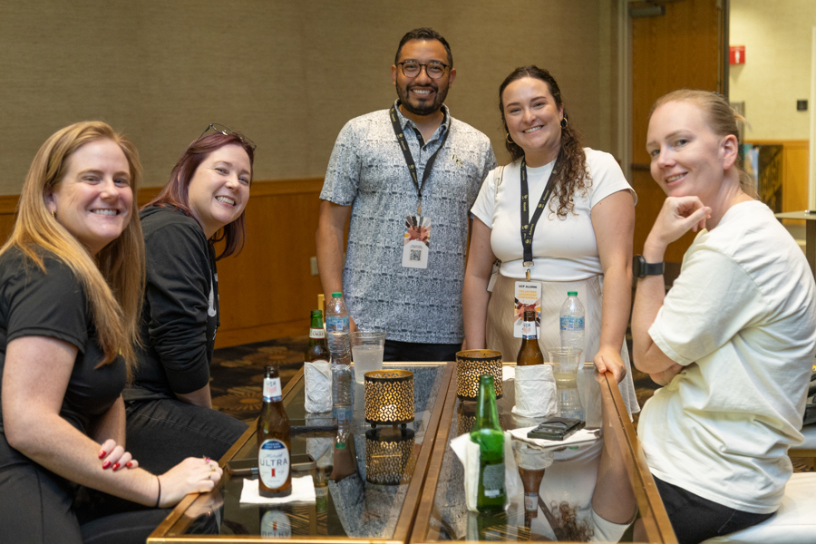 Five adults smiling at a social event, gathered around a glass table with drinks.
