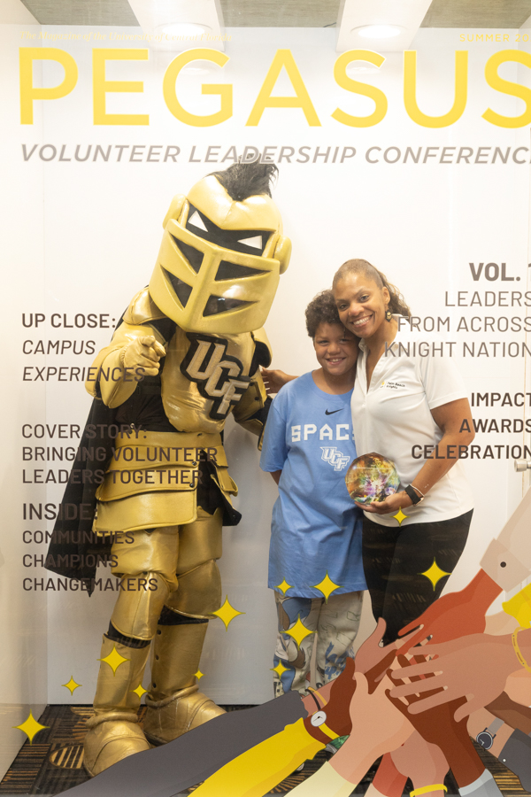 Gold UC mascot posing with a smiling boy in a blue SPACE shirt and a woman at a Pegasus Volunteer Leadership Conference backdrop.
