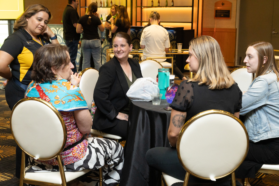 Five women sit around a tall round table at a social event, chatting and smiling with drinks on the table; a bar and others in the background.