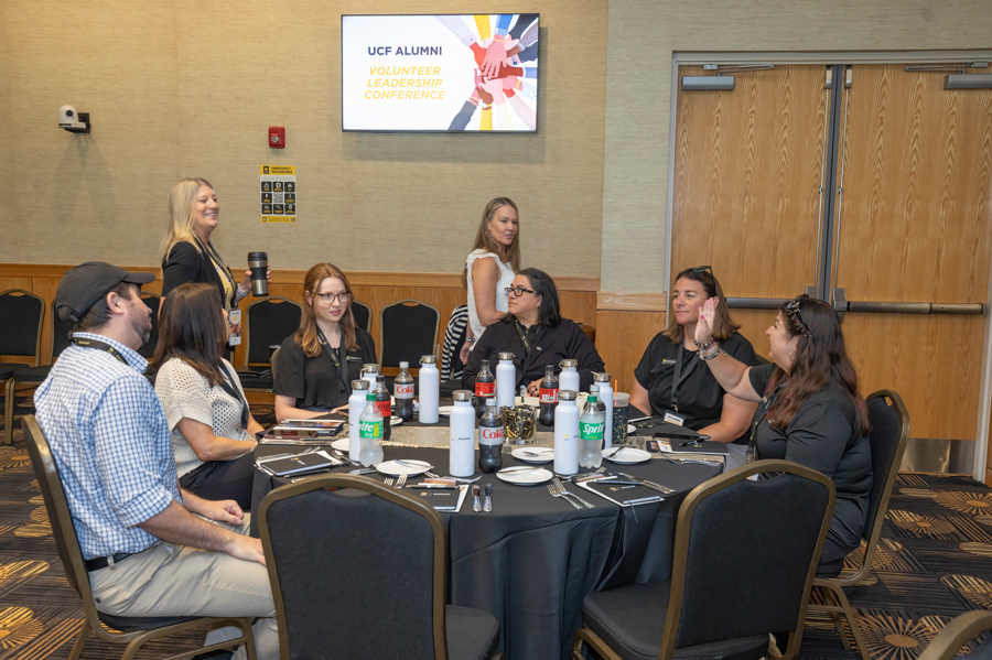 Group of eight adults seated around a round table in a conference room during a UCF Alumni Volunteer Leadership Conference; beverages on the table and a screen with the event title in the background.