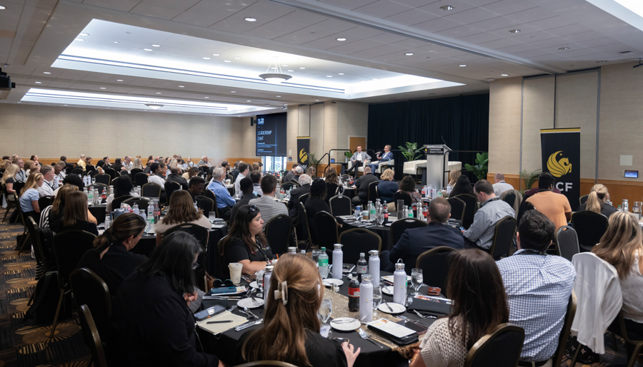 Conference banquet hall with attendees seated at round tables facing a stage where speakers present on a panel.