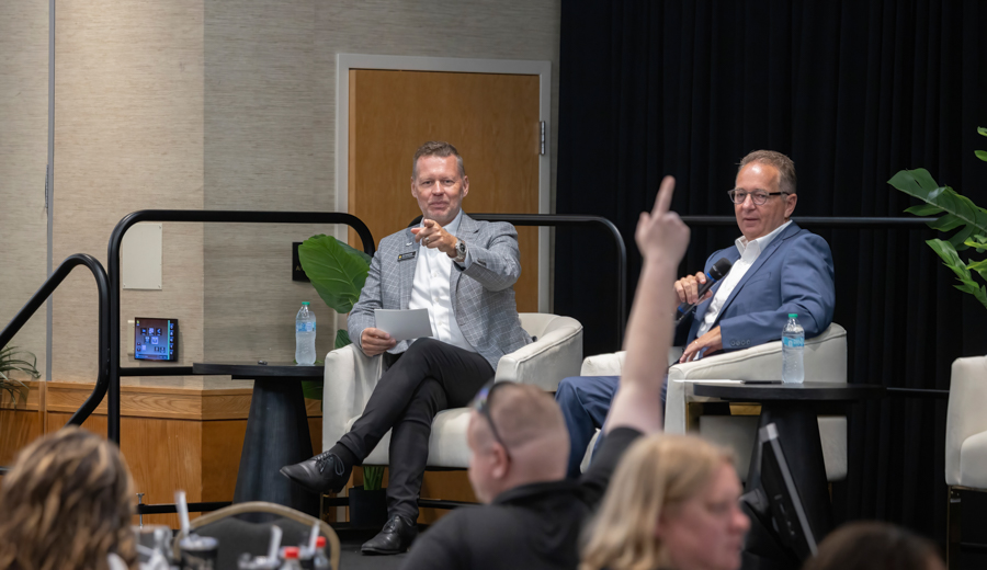 Two men sit on a stage in a panel discussion; one gestures toward the audience while holding notes, the other speaks into a microphone with a water bottle nearby.