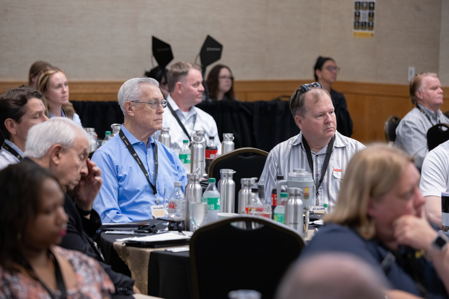 Group of professionals seated at round tables in a conference room, listening to a presentation with water bottles and notepads on the tables.