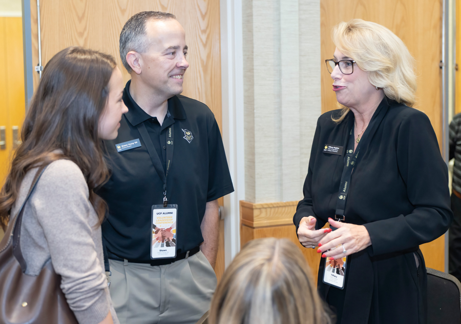 Three conference attendees standing in a hallway, two in black uniforms with name badges, chatting with a guest in a beige sweater.