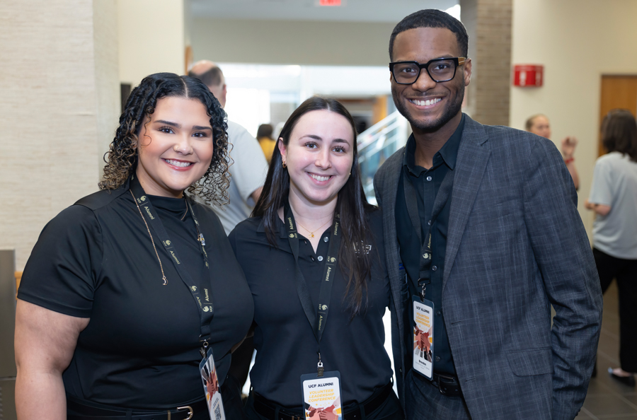 Three smiling conference attendees stand together in a bright lobby, wearing lanyards and name badges.