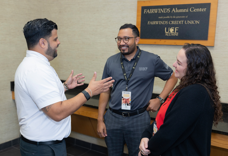 Three people chatting in a lobby by a Fairwinds Alumni Center plaque; man in a white polo gestures as the others listen and smile.