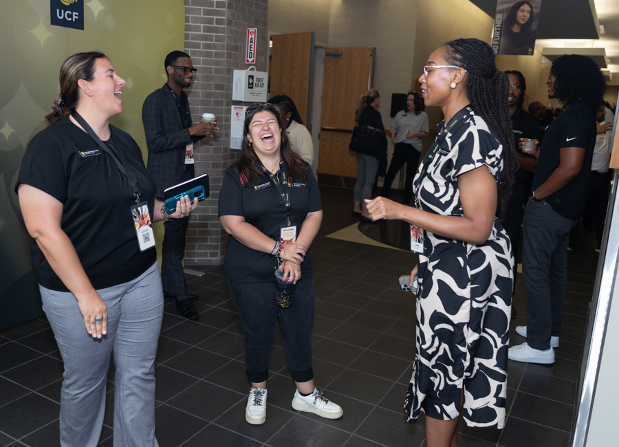 Three staff members laugh and chat in a campus hallway; UCF branding visible on the wall nearby.