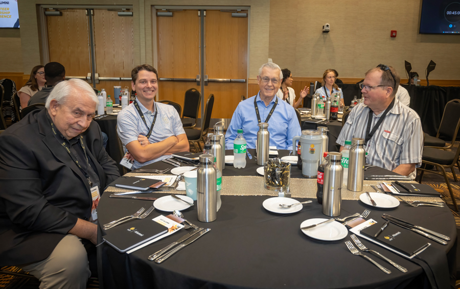 Four men in a conference dining room sit around a round table, smiling at the camera with name badges and water bottles nearby.