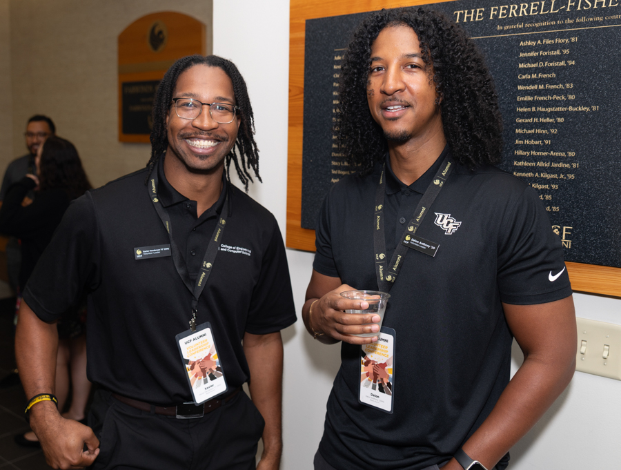 Two men in black polo shirts with lanyards smile at the camera at an indoor event venue.