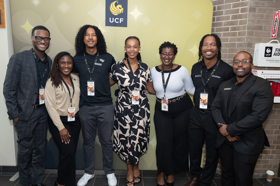 Eight people posing for a group photo in front of a UCF backdrop, all wearing event badges.