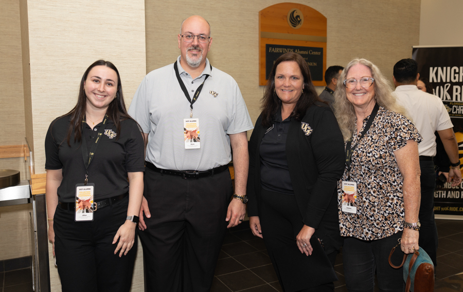 Four adults pose for a photo in a campus alumni center lobby, wearing event badges and smiling at the camera.
