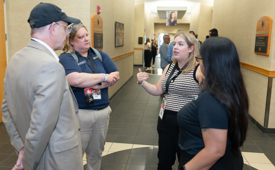 Four people stand in a hallway having a discussion; a man in a beige suit and cap chats with two women wearing lanyards while another woman faces them.