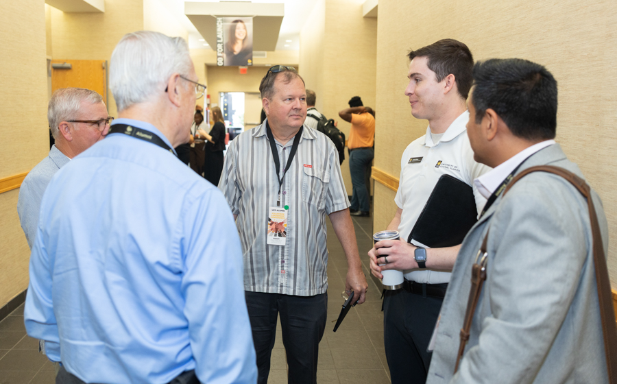 Group of conference attendees in a hallway talking, wearing lanyards and badges; one man holds a folder and another a coffee cup.