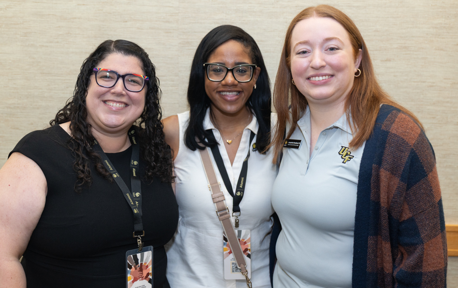 Three smiling women posing together, each wearing lanyards and badges, inside a neutral hallway.