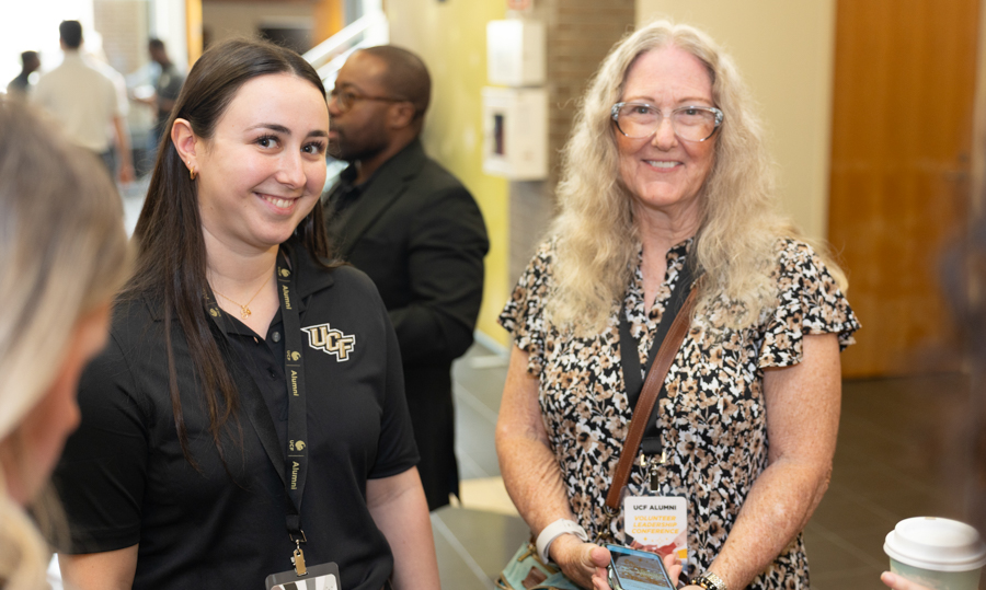Smiling young woman in a UCF polo and lanyard stands beside an older woman with glasses at an indoor event hallway.