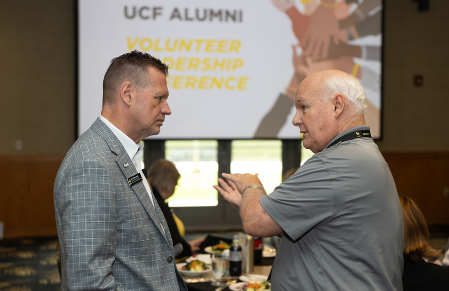 Two men in a conference room talk near tables with lunch, against a backdrop showing 'UCF Alumni Volunteer Leadership Conference'.