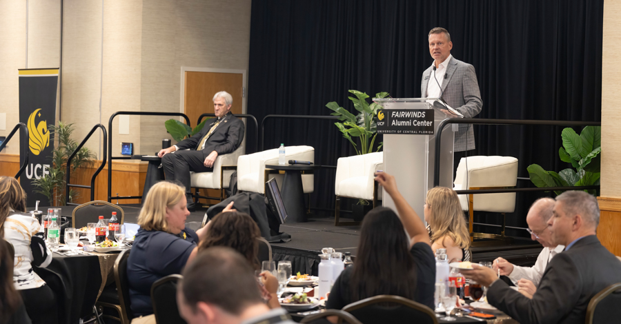 Speaker at a lectern delivering a talk on stage to a seated audience at a formal conference dinner.