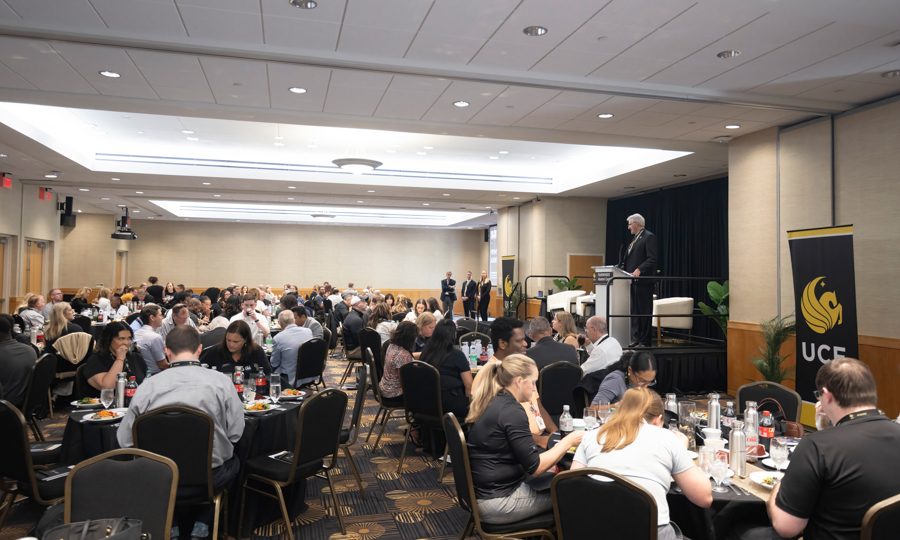 Banquet hall with round tables and a speaker at a podium on stage.