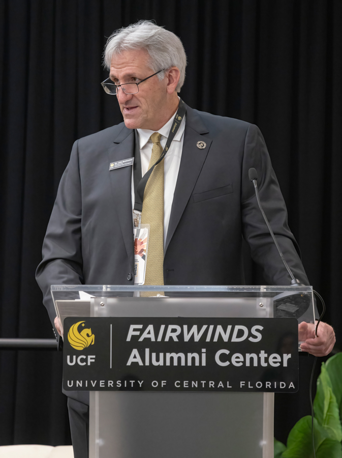Man in a suit giving a speech at a podium labeled 'FAIRWINDS Alumni Center' at UCF, wearing a lanyard and glasses.