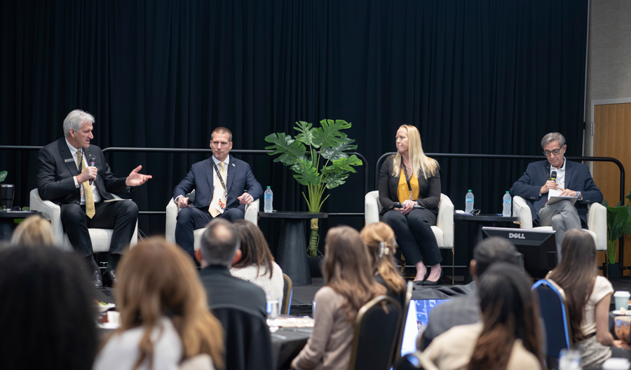 Panel discussion with four speakers seated on a stage in a conference room, audience watching from rows of chairs.