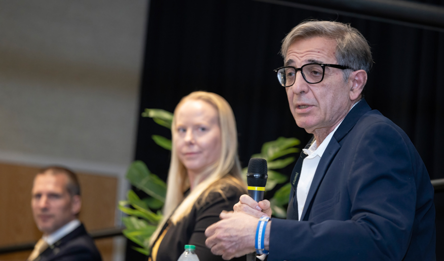 Man in glasses speaks into a handheld microphone at a panel, with two people seated behind him in a conference setting.