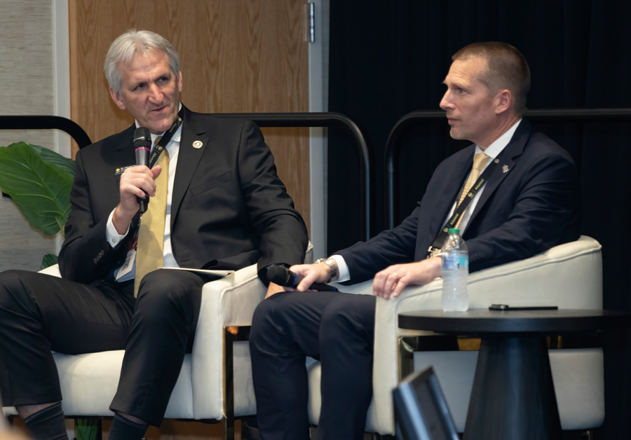 Two men in dark suits seated on a stage, each with a microphone, engaged in a panel discussion.