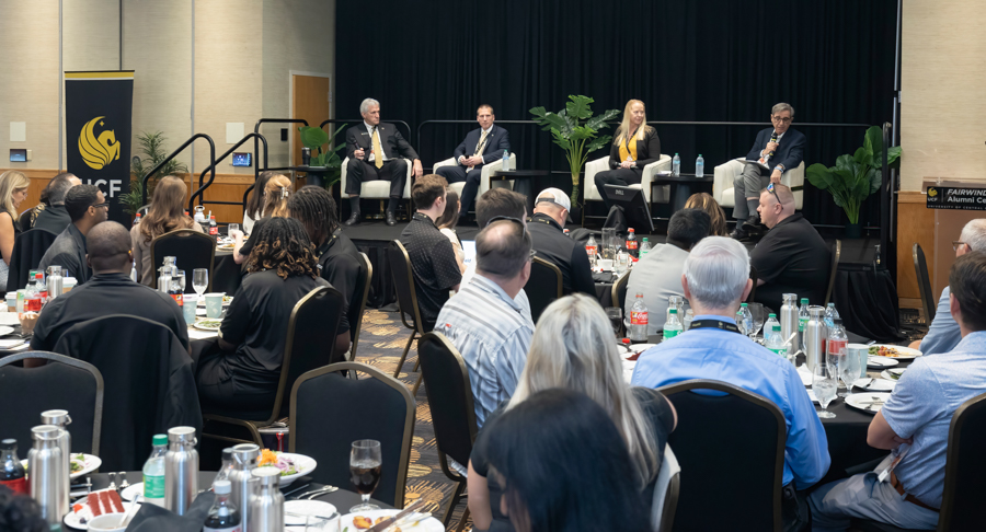 Panel discussion on a stage with four speakers in business attire, facing an audience at round tables with meals and drinks in a conference room.