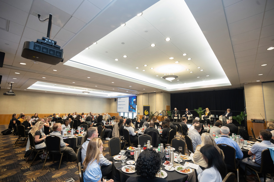 Large conference room filled with round tables and attendees listening to a panel on stage, with a projector screen in the background.