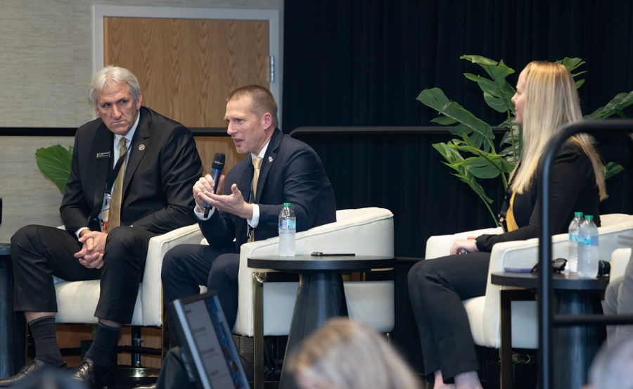 Panel discussion with three adults seated in white chairs; the center man speaks into a handheld microphone while the others listen. 200-? characters