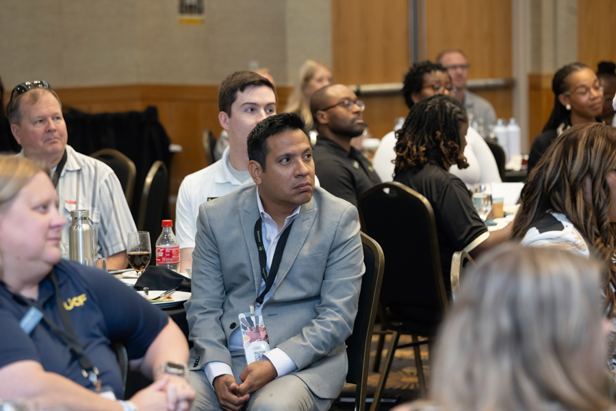 Man in a light grey suit and lanyard seated at a round-table conference, attentively listening among diverse attendees with drinks on the table.