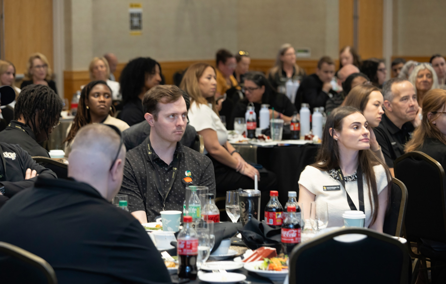 Audience seated at round tables in a conference room, attentively watching a presentation with food and beverages on the tables.