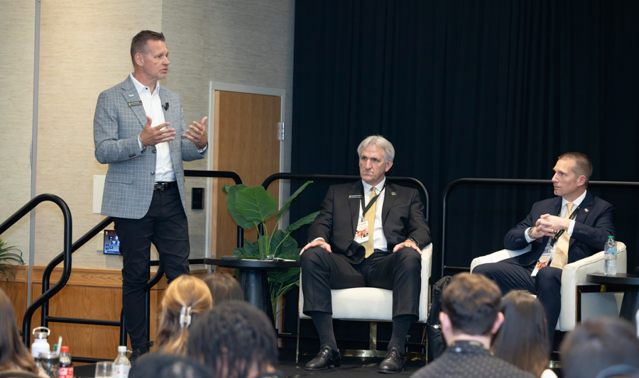 Man stands at a microphone, speaking to an audience while two men in suits listen onstage during a panel discussion in a conference room.