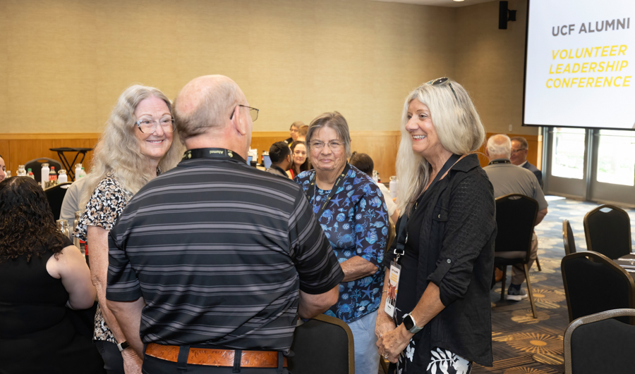Group of four adults chatting and smiling in a conference room, with attendees at tables and a screen reading UCF Alumni Volunteer Leadership Conference in the background.