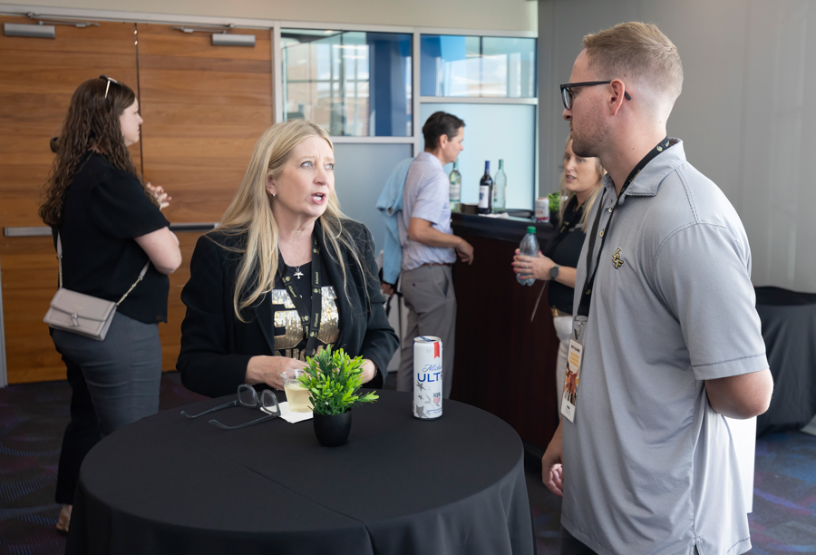 Two people chat at a high-top table during a networking event, with a small potted plant and a can of Ultra beside them in a lounge area.