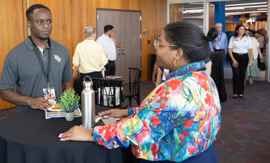 Man in a UF polo with a badge talks to a guest across a round table at a conference, with a plant and water bottle on the table.